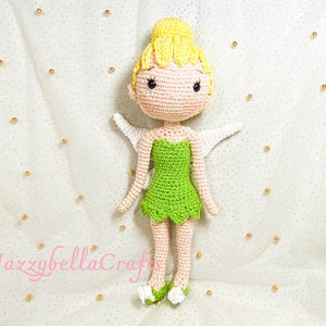 Puede incluir: Una muñeca de ganchillo de Campanilla con vestido verde, pelo amarillo y alas blancas. La muñeca está sentada sobre un fondo blanco con cuentas doradas.