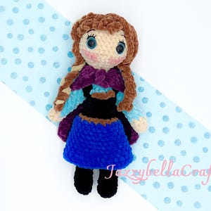 May include: A crocheted doll with brown hair, blue eyes, and a purple and blue dress. The doll is wearing black shoes and is standing on a blue and white polka dot background.