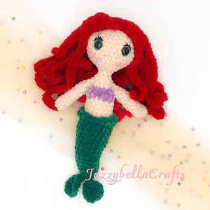 Può includere: Una bambola di sirena all'uncinetto con capelli rossi, un reggiseno a conchiglia viola e una coda verde. La bambola è sdraiata su un tessuto di tulle beige con paillettes colorate.