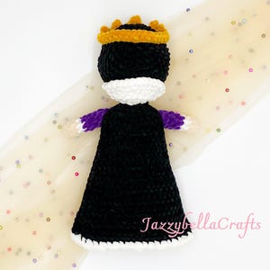 Pode incluir: Boneca de croch&ecirc; feita &agrave; m&atilde;o em forma de rainha, com vestido preto, gola branca e coroa dourada. A boneca tem mangas roxas e est&aacute; sobre um fundo claro com lantejoulas espalhadas.