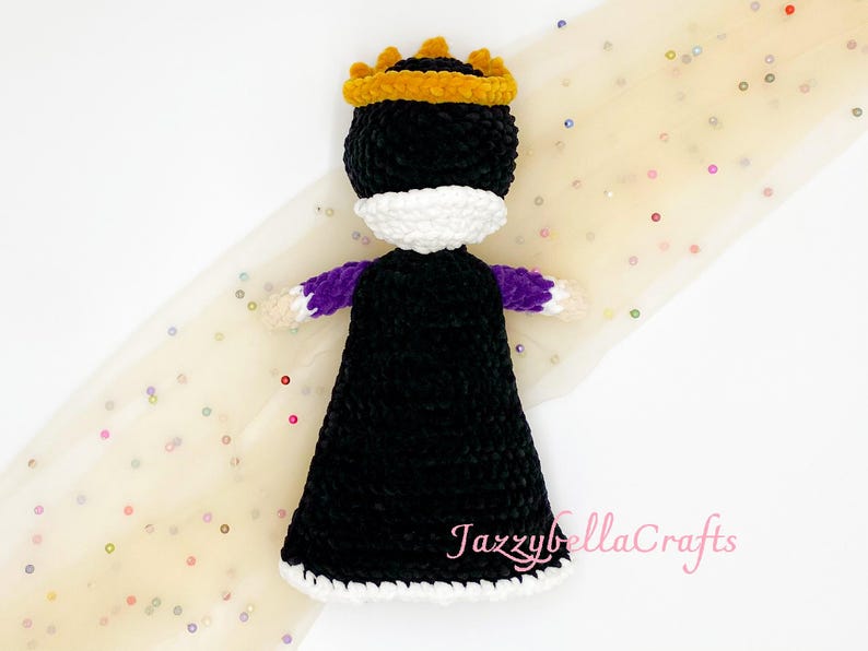 Pode incluir: Boneca de croch&ecirc; feita &agrave; m&atilde;o em forma de rainha, com vestido preto, gola branca e coroa dourada. A boneca tem mangas roxas e est&aacute; sobre um fundo claro com lantejoulas espalhadas.