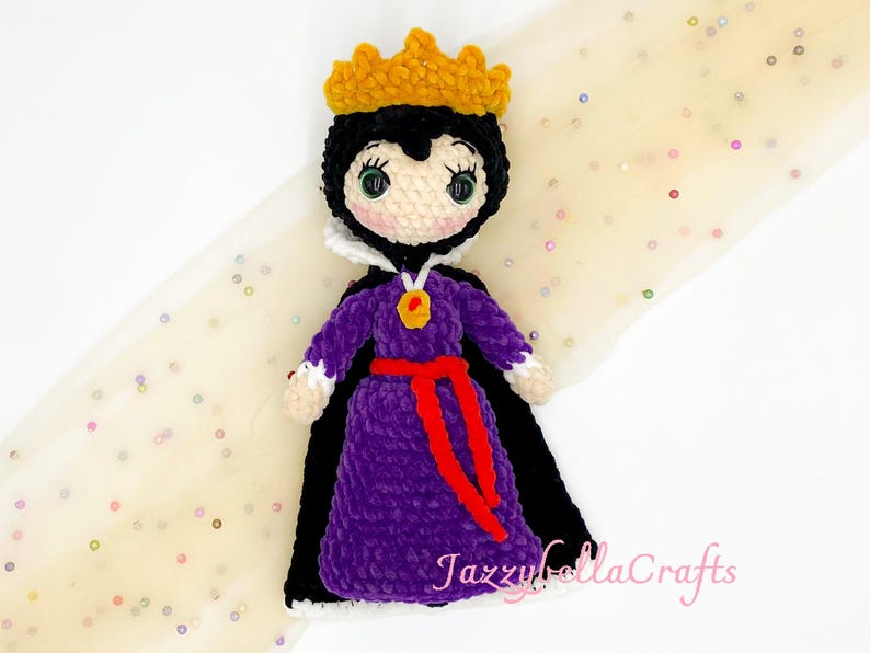 Pode incluir: Boneca de croch&ecirc; de uma rainha usando uma coroa de ouro, capa preta e vestido roxo com cinto vermelho. A boneca tem uma gola branca e um pingente de ouro. O rosto da boneca &eacute; branco com cabelo preto e olhos verdes.