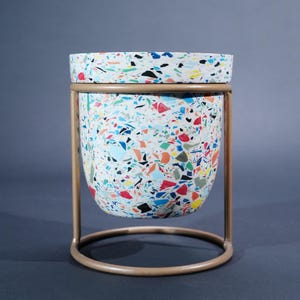 May include: A decorative planter with a terrazzo-style design, featuring a white base speckled with colourful chips of red, blue, yellow, and black. The planter sits in a gold-coloured metal stand, adding a touch of modern style.