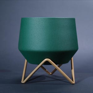 Peut inclure: Un pot de fleur vert foncé avec une surface texturée, posé sur un support en métal doré. Le pot a une forme géométrique avec des côtés inclinés. Le support a une base triangulaire. Le design est moderne et minimaliste.