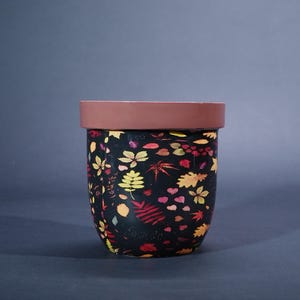 May include: A black ceramic plant pot with a brown rim, decorated with a colourful leaf pattern. The leaves are in shades of yellow, red, and orange, creating an autumnal design. The pot is a decorative item for indoor or outdoor use.