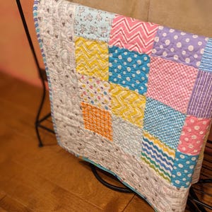 Può includere: Una colorata trapunta patchwork con una varietà di motivi e colori, tra cui pois, zig-zag e quadrati. La trapunta è esposta su un supporto in metallo nero. Il bordo della trapunta è rifinito con un bordo blu chiaro.