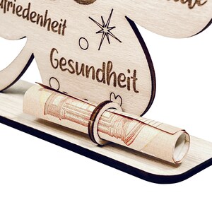 May include: Wooden money holder shaped like a four-leaf clover with the words "Friedenheit" and "Gesundheit" engraved on it. A rolled-up banknote is placed in the holder.