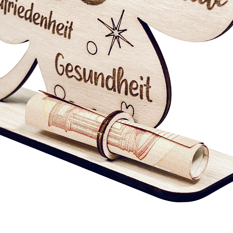 May include: Wooden money holder shaped like a four-leaf clover with the words "Friedenheit" and "Gesundheit" engraved on it. A rolled-up banknote is placed in the holder.