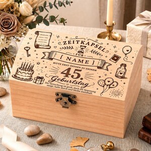 Puede incluir: Una caja de madera de color claro con tapa con bisagras y cierre metálico. La tapa está decorada con ilustraciones grabadas de una tarta, globos y texto en alemán, incluyendo "ZEITKAPSEL" y "45. Geburtstag."