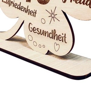 May include: A wooden clover-shaped sign with the words "Gl&uuml;ck", "Liebe", "Friedenheit", and "Gesundheit" engraved on it. The sign has a small stand on the bottom.