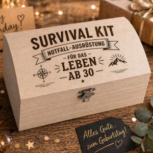 Könnte beinhalten: Eine hellfarbene Holzkiste mit den Worten "SURVIVAL KIT" und "NOTFALL-AUSRÜSTUNG" auf dem Deckel. Die Kiste zeigt auch einen Kompass, eine Berg-Grafik und den Text "FÜR DAS LEBEN AB 30". Eine kleine schwarze Karte mit goldenem Text lautet "Alles Gute zum Geburtstag!"