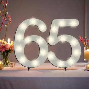 Op de afbeelding: Witte houten cijfer 65 met witte lampjes, staand op een witte tafel met een wit tafelkleed. De cijfers zijn verlicht en zijn een populaire decoratie voor verjaardagen en jubileums.