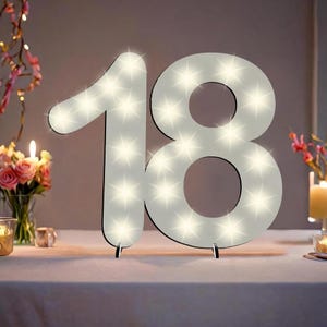 Può includere: Numero 18 in legno bianco illuminato con luci LED bianche. Il numero è su un tavolo bianco.