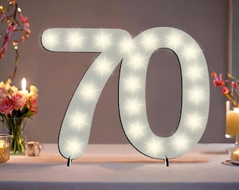 May include: White wooden number 70 with white lights, standing on a white table. The number is lit up and is a decorative piece for a party or celebration.