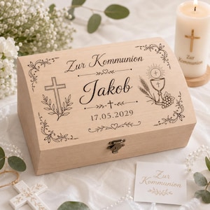 Può includere: Una scatola di legno color chiaro con coperchio incernierato, incisa con le parole "Zur Kommunion" e il nome "Jakob" insieme alla data 17.05.2029. La scatola presenta una croce, decorazioni floreali e un calice. È visibile una candela con una croce.