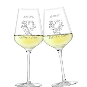 Puede incluir: Dos copas de vino transparentes llenas de vino blanco. Cada copa está grabada con un diseño de corazón y rosa, la fecha 25.01.2023 y los nombres "Celine & Alex". Las copas están sobre un fondo blanco.