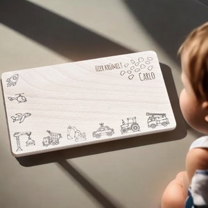 Könnte beinhalten: Ein weißes Holzbrett mit dem Text "Hier krümelt Carlo" und verschiedenen Fahrzeugillustrationen, darunter eine Rakete, ein Hubschrauber, ein Flugzeug, ein Kran, ein Kipplaster, ein Bagger, ein Auto, ein Traktor und ein Feuerwehrauto.