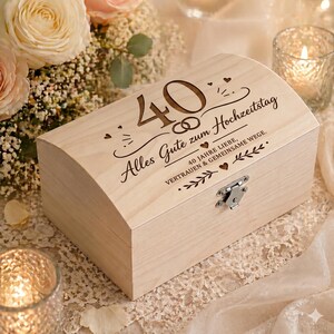 Könnte beinhalten: Eine hellfarbene Holzkiste mit gewölbtem Deckel, mit graviertem Text und einem Metallverschluss. Der Text enthält die Zahl "40" und die Worte "Alles Gute zum Hochzeitstag". Die Kiste ist von Blumen und Kerzen umgeben.