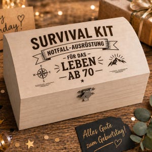 Puede incluir: Una caja de madera de color claro con las palabras "SURVIVAL KIT" y "NOTFALL-AUSRÜSTUNG" impresas en la tapa. La caja también presenta el texto "FÜR DAS - LEBEN - AB 70-" y un diseño de rosa de los vientos. Se ve una pequeña tarjeta negra con texto dorado que dice "Alles Gute zum Geburtstag!"