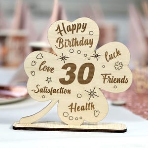 May include: Wooden clover-shaped sign with the text "Happy Birthday 30" and the words "love, satisfaction, health, luck, friends" around the number 30.