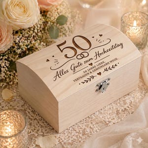 Op de afbeelding: Een lichtgekleurde houten kist met een gebogen deksel. De kist is gegraveerd met het nummer "50", trouwringen en de woorden "Alles Gute zum Hochzeitstag". Een metalen sluiting zekert de kist.