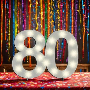 Op de afbeelding: Witte verlichte cijfer 80 met een witte achtergrond en een zwarte omtrek. Het cijfer staat op een tafel met een rood-wit tafelkleed. De achtergrond is een muur met kleurrijke slingers.