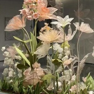 Puede incluir: Una vitrina de cristal llena de delicadas flores de papel en colores pastel. Las flores est&aacute;n dispuestas en una variedad de formas y tama&ntilde;os, creando un ambiente caprichoso y rom&aacute;ntico.