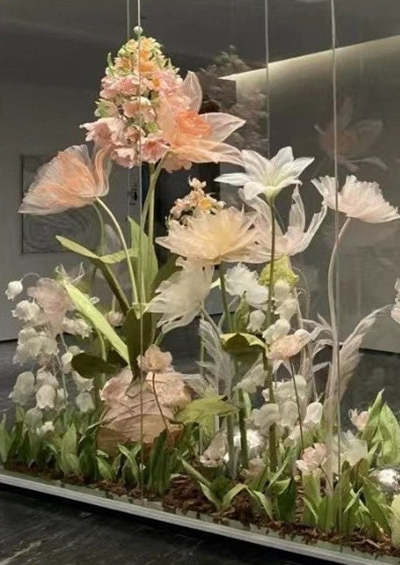 Puede incluir: Una vitrina de cristal llena de delicadas flores de papel en colores pastel. Las flores est&aacute;n dispuestas en una variedad de formas y tama&ntilde;os, creando un ambiente caprichoso y rom&aacute;ntico.