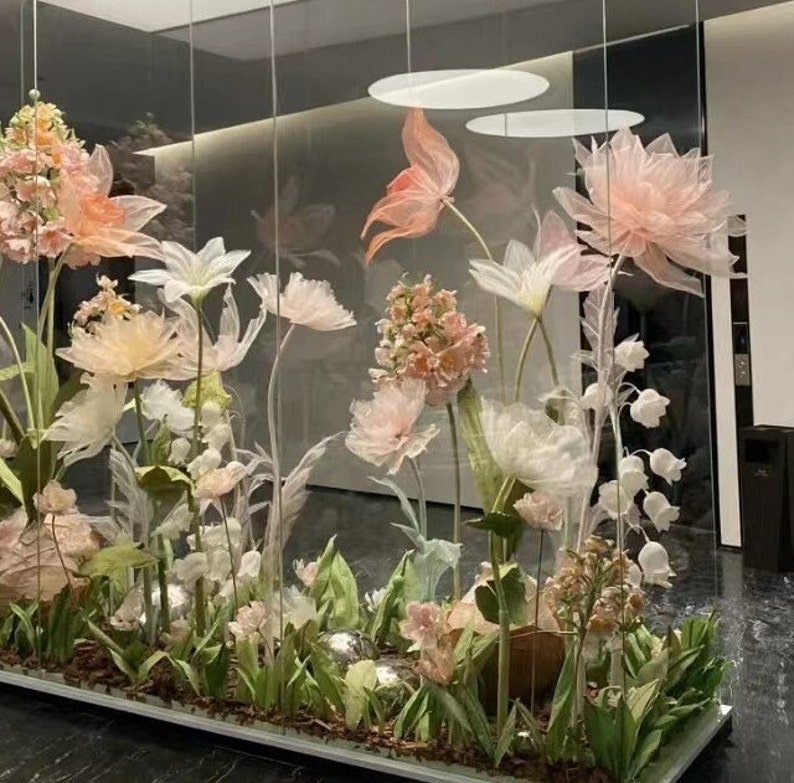 Puede incluir: Una vitrina de cristal llena de delicadas flores de papel en colores pastel. Las flores est&aacute;n dispuestas en una variedad de formas y tama&ntilde;os, creando un ambiente caprichoso y rom&aacute;ntico.