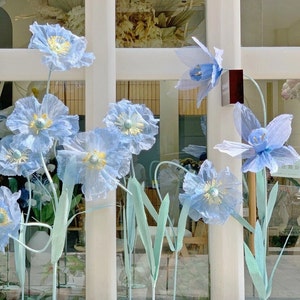 Puede incluir: Una exhibici&oacute;n de flores de papel azul con tallos verdes frente a una ventana. Las flores est&aacute;n dispuestas en un jarr&oacute;n y est&aacute;n en diferentes etapas de floraci&oacute;n.