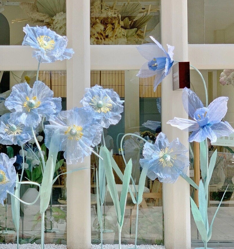Puede incluir: Una exhibici&oacute;n de flores de papel azul con tallos verdes frente a una ventana. Las flores est&aacute;n dispuestas en un jarr&oacute;n y est&aacute;n en diferentes etapas de floraci&oacute;n.
