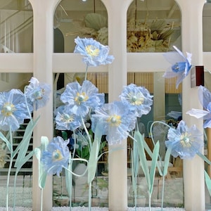 Puede incluir: Un escaparate de tienda con una variedad de flores de papel azul. Las flores est&aacute;n dispuestas en un racimo y est&aacute;n unidas a tallos que se extienden desde la parte inferior de la ventana. Las flores son de diferentes tama&ntilde;os y formas, y algunas tienen una textura delicada y rizada.
