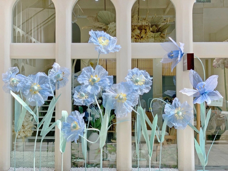 Puede incluir: Un escaparate de tienda con una variedad de flores de papel azul. Las flores est&aacute;n dispuestas en un racimo y est&aacute;n unidas a tallos que se extienden desde la parte inferior de la ventana. Las flores son de diferentes tama&ntilde;os y formas, y algunas tienen una textura delicada y rizada.