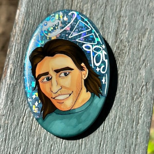 May include: An oval-shaped pin featuring a cartoon illustration of a person with brown hair and a teal shirt. The background is a blue holographic glitter finish with white line art of a small creature and the text 985 written in white.
