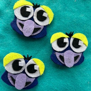 May include: Three felt owl-like faces with large white and black eyes, yellow eyebrows, and purple beaks. The faces are primarily blue with black details, arranged on a teal background.