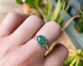 May include: A silver ring featuring a faceted, oval-shaped green gemstone. The gemstone is set in a bezel setting with a scalloped edge. The ring is shown on a person's finger, with a blurred background of greenery.