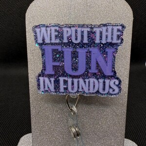 Könnte beinhalten: Ein Ausweis-Rollhalter mit dem Text "WE PUT THE FUN IN FUNDUS" in Lila und glitzerndem Blau. Der Ausweis-Rollhalter ist an einem transparenten Kunststoffhalter und einem silbernen Metallclip befestigt.