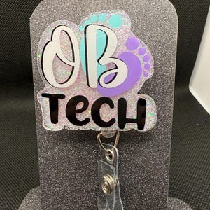 May include: A glittery badge reel with the text "OB TECH" in black and white. The design includes a baby footprint in teal and purple. The badge reel has a clear plastic strap and a metal clip.