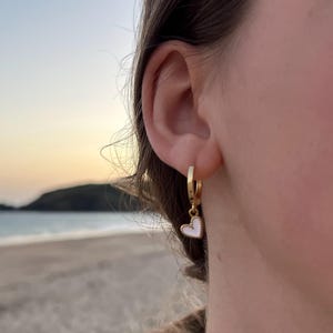 Może przedstawiać: Złoty kolczyk koło z białą zawieszką w kształcie serca. Kolczyk jest noszony na uchu osoby. Tło to scena na plaży ze wschodem słońca.