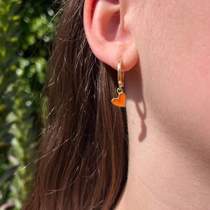 May include: Gold hoop earring featuring a small orange heart charm. The heart is a vibrant, solid orange. The earring is worn on an ear, with long brown hair visible.