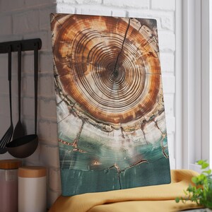 Puede incluir: Tabla de cortar rectangular con una sección transversal detallada de un tronco de árbol. La veta de la madera presenta círculos concéntricos en tonos marrón y beige, con una sección inferior turquesa y blanca. La tabla descansa sobre un paño amarillo.