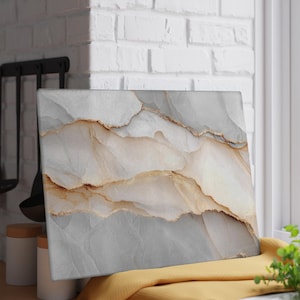 May include: A rectangular chopping board with a marble design. The board features a blend of grey, white, and gold tones, creating a layered effect. The board is displayed on a yellow cloth near a window.
