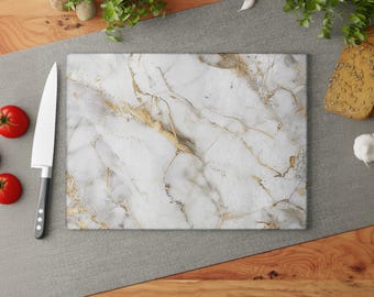 May include: Rectangular white marble cutting board with gold veining. A large chef's knife and three red tomatoes are to the left. The board is on a gray surface with bread and herbs in the background.