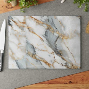 May include: Rectangular marble-patterned cutting board with gold veining. A stainless steel knife with a black handle rests beside the board. Two red tomatoes are visible, along with bread and herbs.