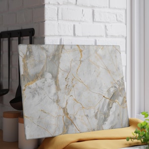 May include: A rectangular cutting board with a marble design. The board features a white and gray background with gold veining. It is resting on a yellow cloth in front of a white brick wall and a window.