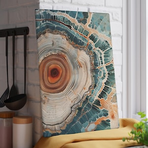 May include: Rectangular wall art with a detailed abstract design. The artwork features concentric circles in shades of brown, beige, and orange, surrounded by teal and white patterns. The piece is displayed on a yellow surface near a window.
