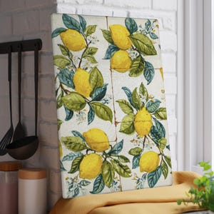 May include: Rectangular cutting board with a lemon design. The board features a repeating pattern of yellow lemons, green leaves, and white blossoms on a cream-colored background. The board is leaning against a white brick wall.