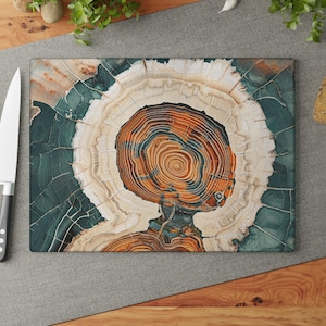 May include: Rectangular cutting board with a tree ring design in shades of orange, white, and teal. A stainless steel knife and fresh red tomatoes are next to the board. The board is on a gray surface.