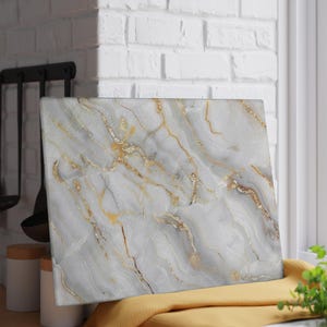 Può includere: Tagliere rettangolare con un design marmorizzato. La tavola presenta uno sfondo bianco e grigio con venature dorate. La tavola è appoggiata su un panno giallo.