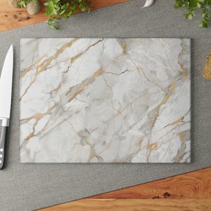 May include: Rectangular white marble cutting board with gold veining. A stainless steel knife with a black handle rests on a gray surface next to the board. Red tomatoes, bread, and garlic are also visible.